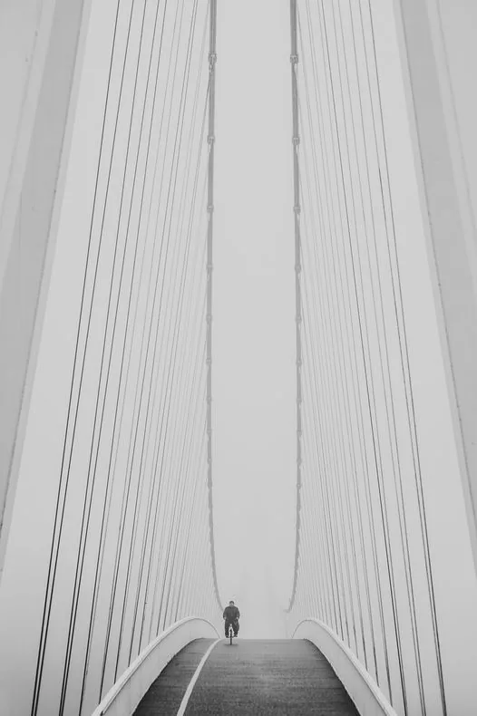 Dramatic monochrome image of a lone cyclist riding over a suspension bridge.