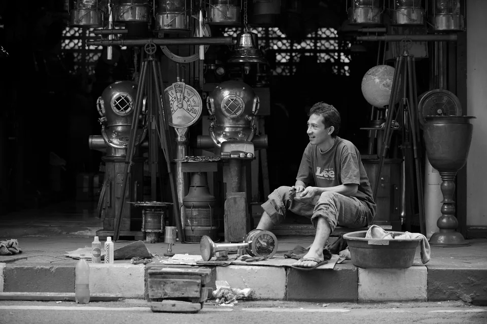 A man sits outside a shop displaying metal pans — photograph by Peter Pickering