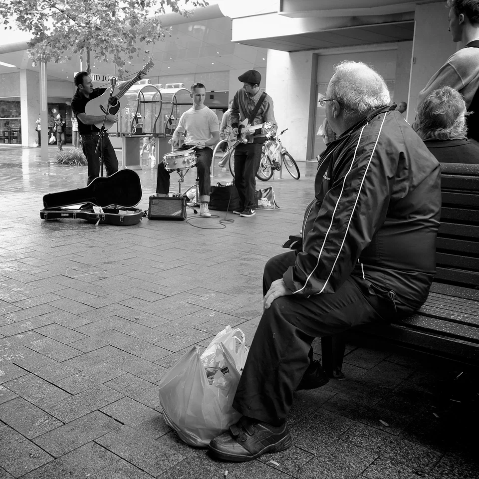 An older listener watches musicians from a street bench — photograph by Peter Pickering