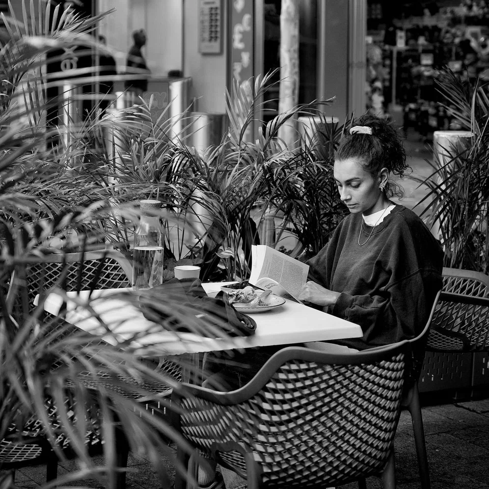 A woman reads at a café table surrounded by foliage — photograph by Peter Pickering