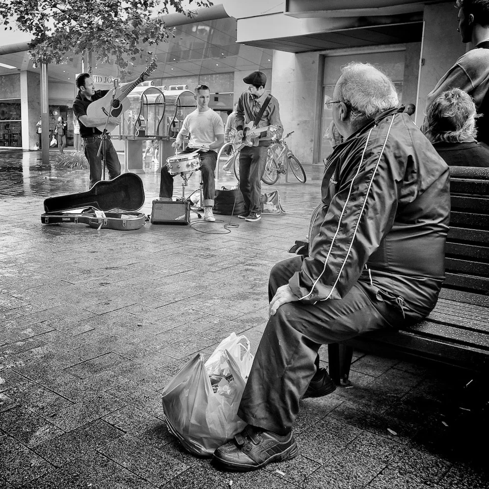 Older man sitting on mall bench listening to busking rock and roll group